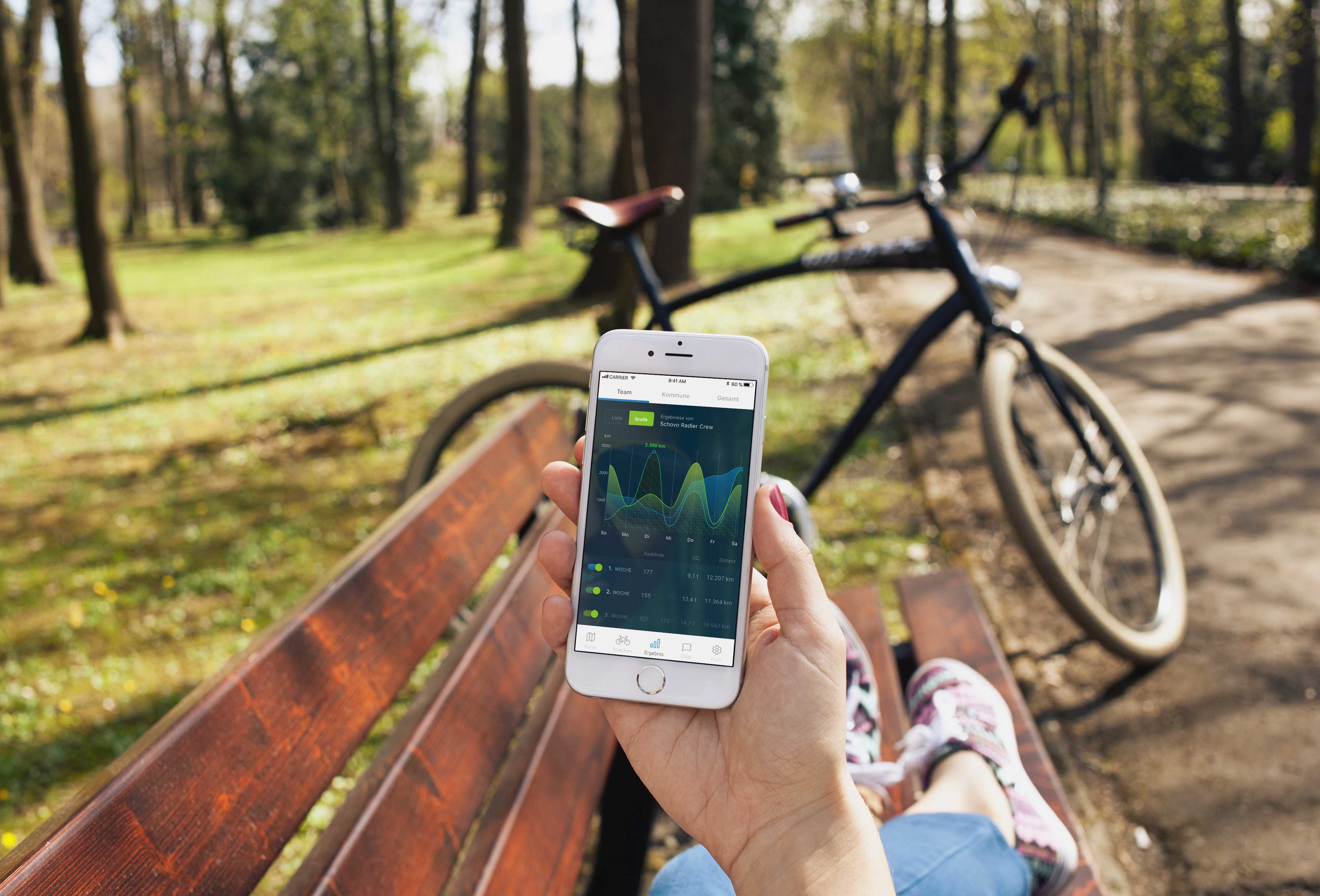 Hand mit Handy und Beine eines Menschen, der auf einer Bank sitzt, im Hintergrund ein Fahrrad und Bäume