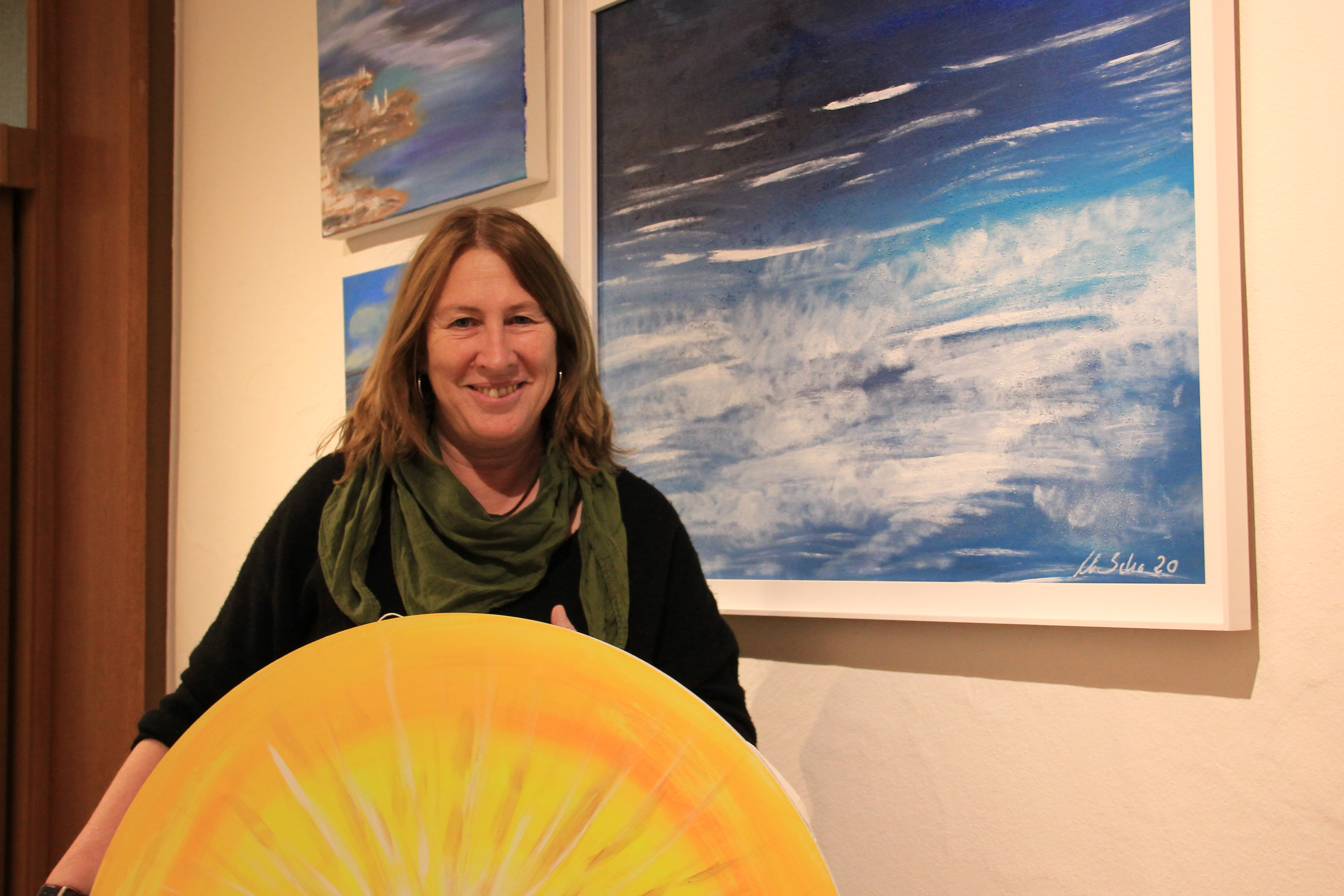 Künstlerin, die ein gelbes Sonnenbild in der Hand hält und vor blauen Meeransichten steht