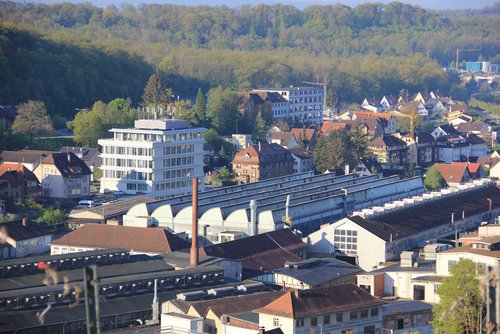 Blick von einer Anhöhe auf eine Fabrikgelände mit verschiedenen Hallen, Gebäuden, Schornstein