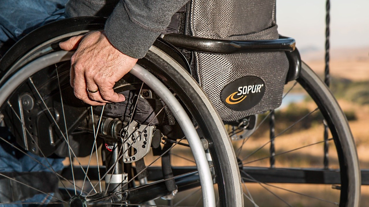 Zoom auf die Räder eines Rollstuhls, auf dem eni Mann sitzt, eine Hand am Rad