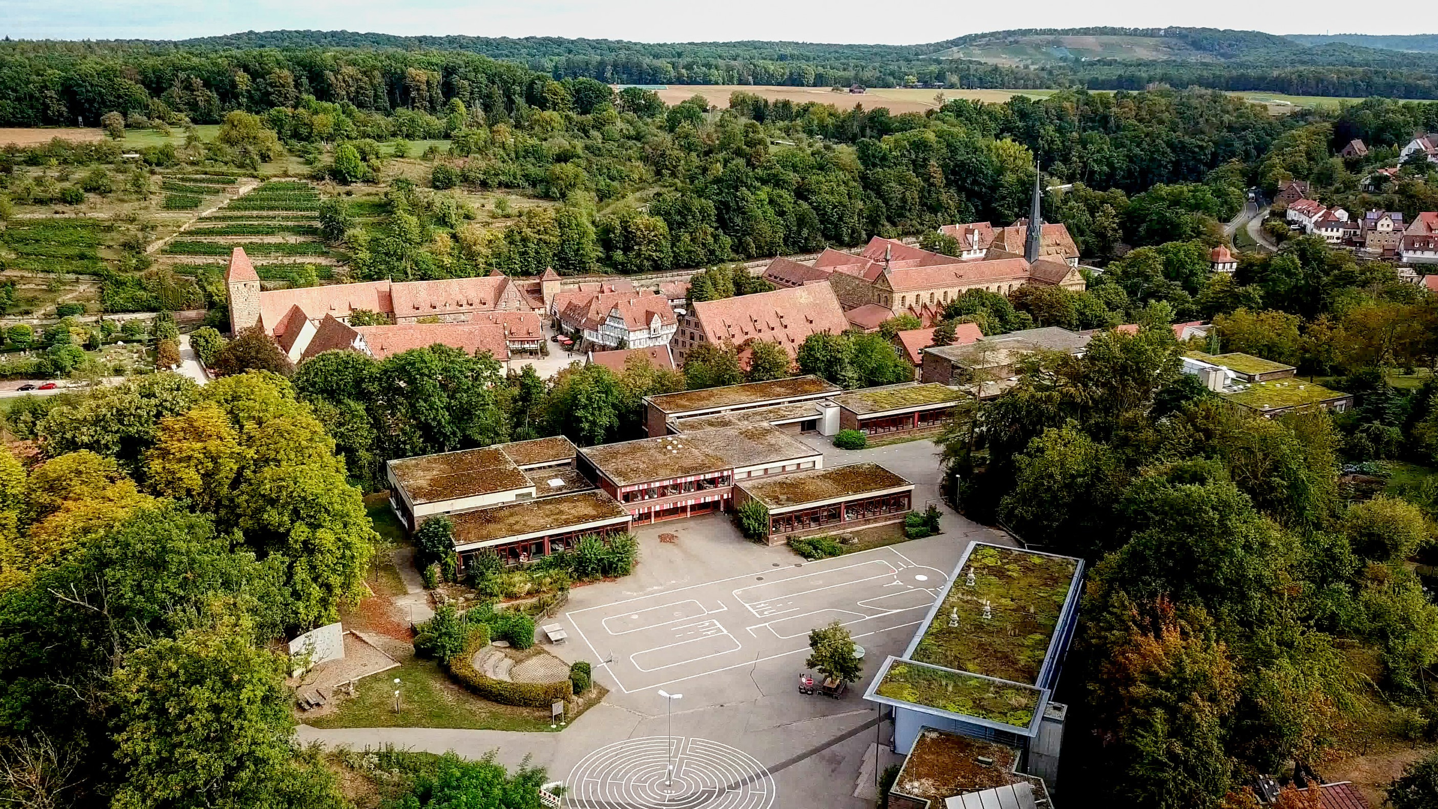 Blick auf ein Ensemble von Schulgebäuden aus Sandstein, den Schulhof und Bäume, im Hintergrund das Kloster