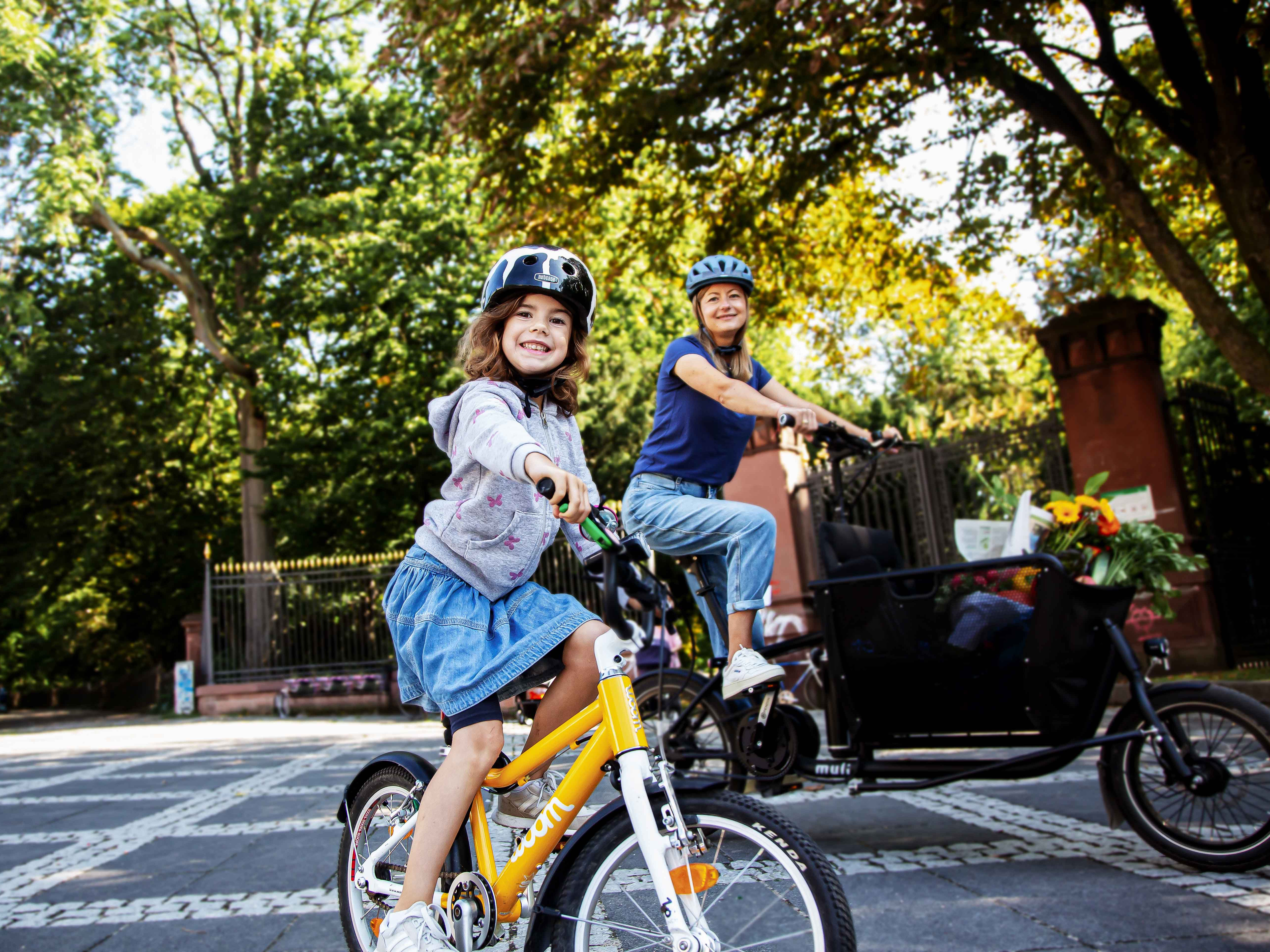 Frau mit Lastenbike und kleines Mädchen auf gelbem Fahrrad vor einem Park