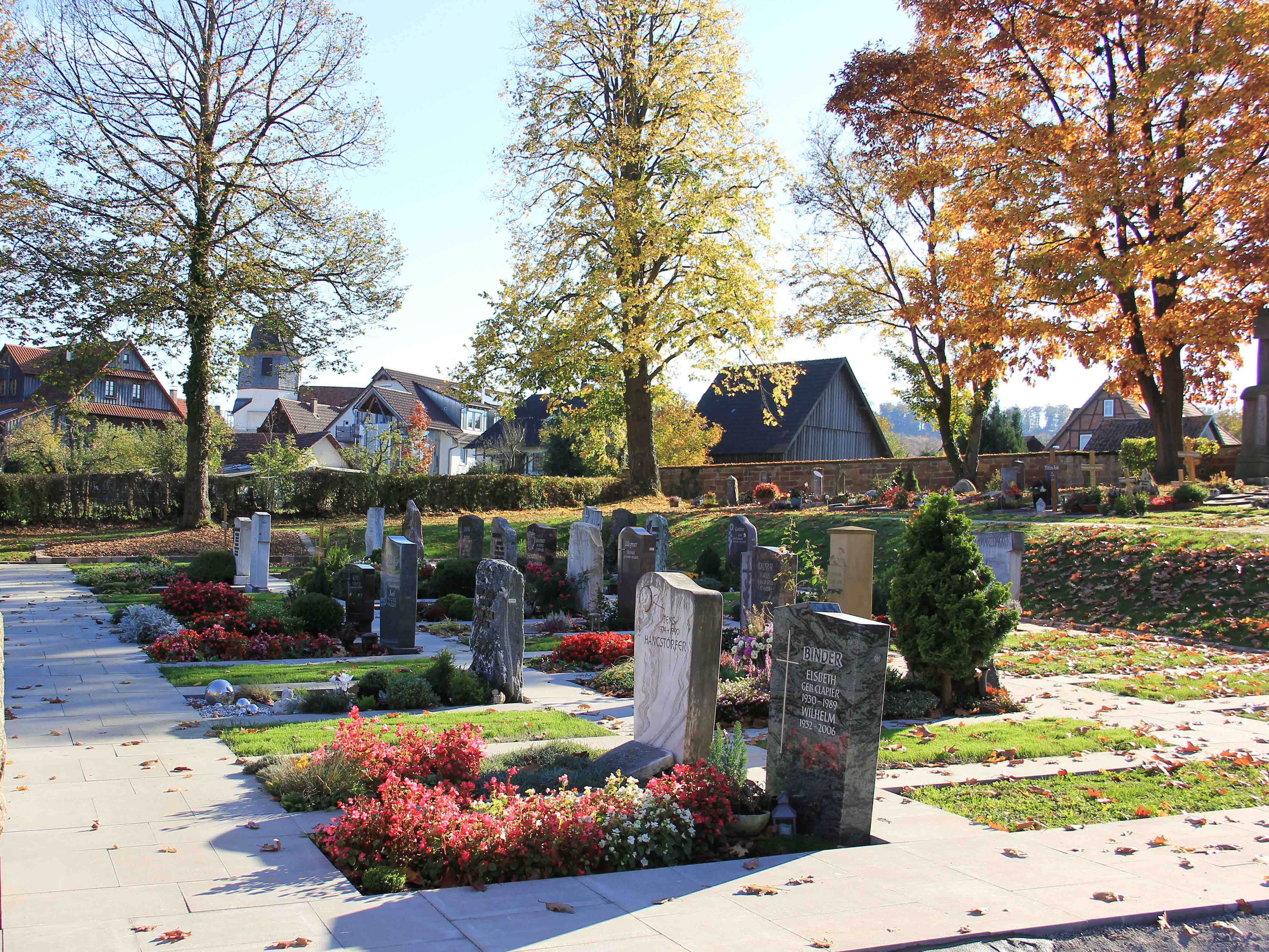 Blumenreich bepflanzte Erdbestattungsgräber mit Grabsteinen, im Hintergrund ist eine Dorfsilhouette zu erkennen